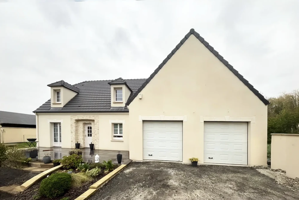 Maison individuelle contemporaine avec façade en enduit beige clair, toiture en tuiles grises et deux lucarnes