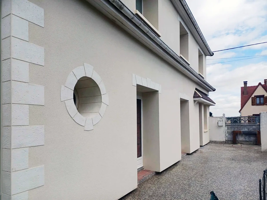 Façade de bâtiment moderne avec mur crépi beige, fenêtre circulaire ornementale à encadrement blanc en saillie, colonnes rectangulaires et porche couvert