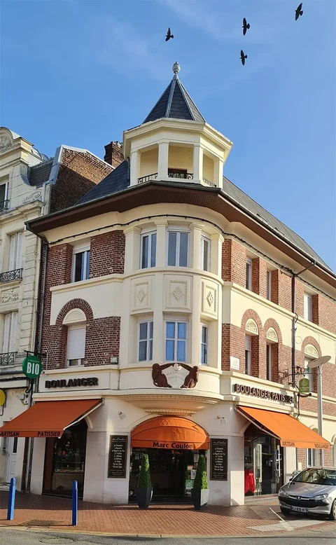 Scène extérieure urbaine montrant un bâtiment commercial d'angle avec façade en briques rouges et éléments architecturaux blancs